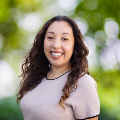 A woman with long, curly hair smiles warmly at the camera. She is wearing a light-colored top with black trim. The background is blurred greenery, suggesting an outdoor setting.