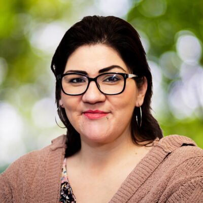 Woman with dark hair and glasses, wearing earrings, a patterned shirt, and a knitted brown cardigan. She is smiling against a backdrop of blurred greenery.