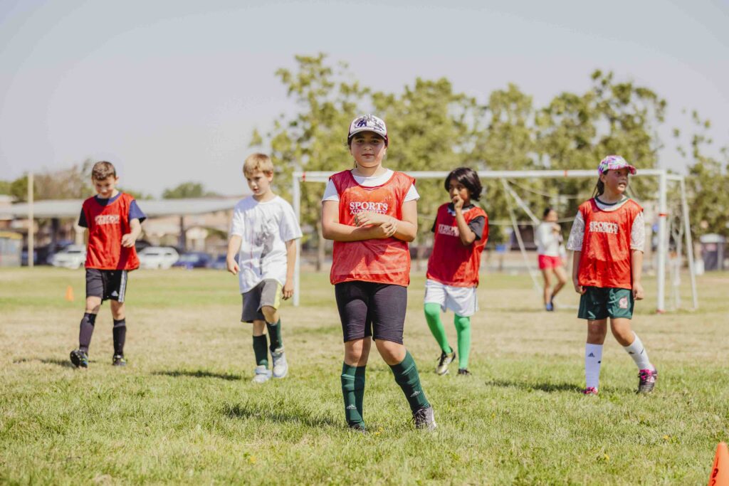 Five children wearing sports jerseys stand on a grassy soccer field, with goalposts and trees in the background. The kids appear to be taking a break, some with hands on hips or arms crossed.
