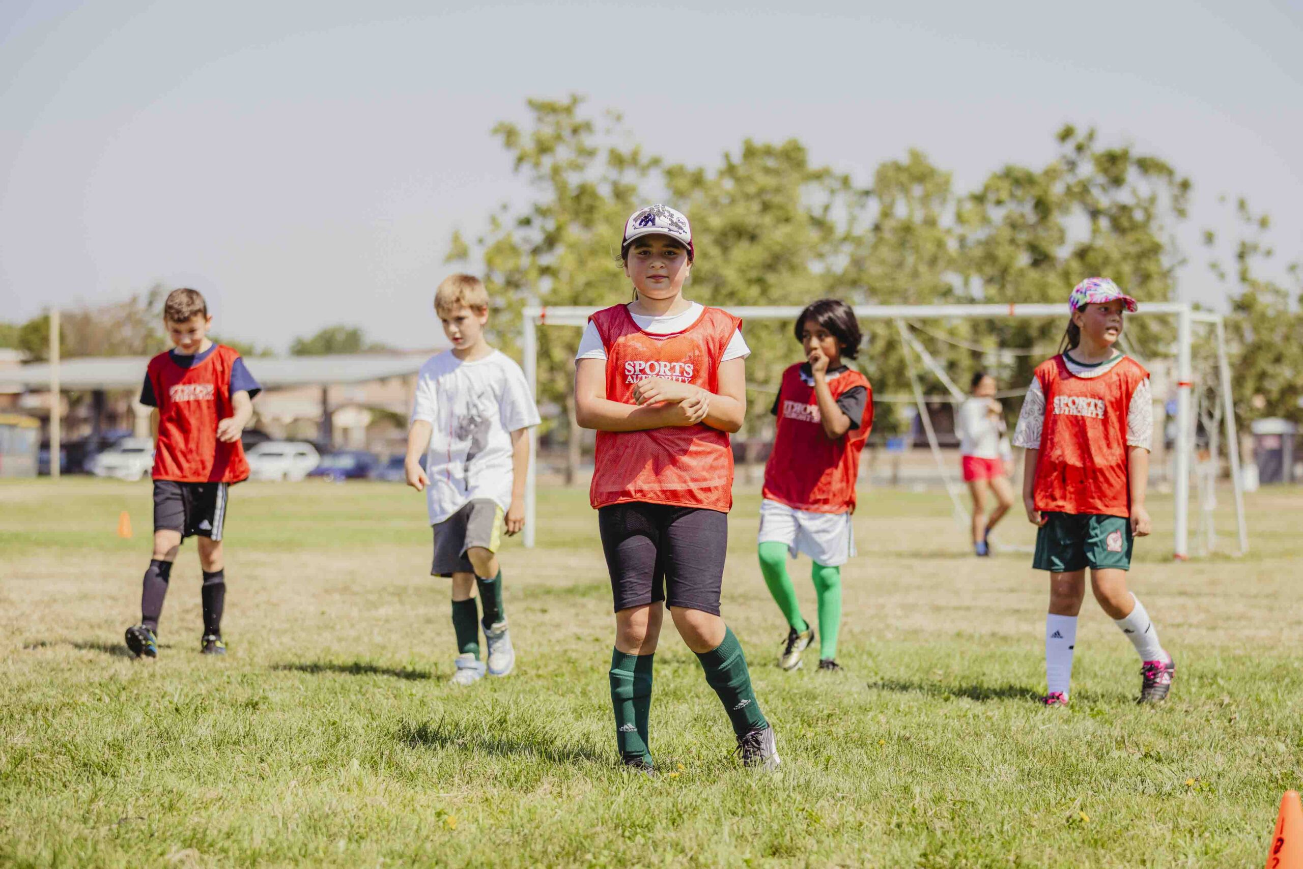 Five children wearing sports jerseys stand on a grassy soccer field, with goalposts and trees in the background. The kids appear to be taking a break, some with hands on hips or arms crossed.