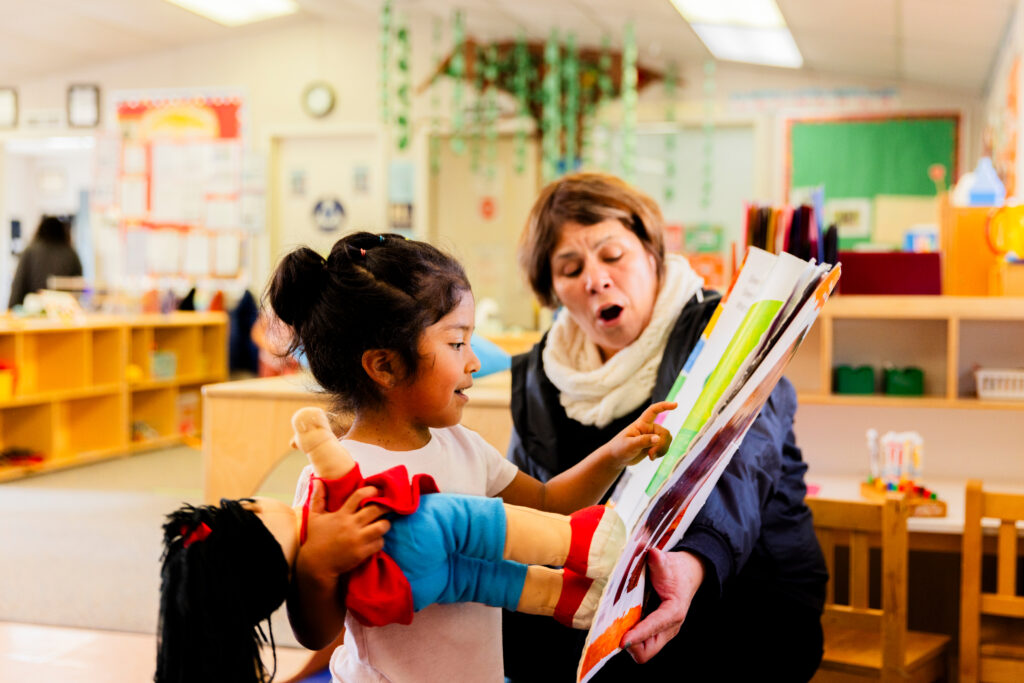 A young girl holding a doll excitedly points at a book while an adult woman beside her reads aloud in a colorful classroom setting.