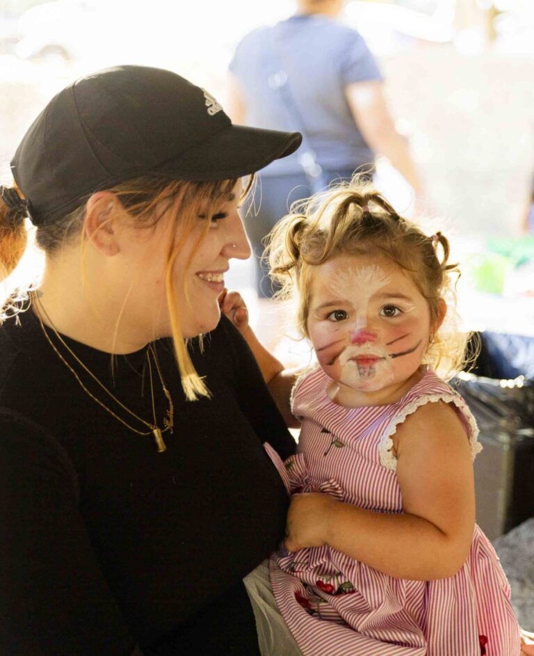 A woman in a black shirt and cap smiles while holding a young girl with face paint resembling a cat, wearing a pink dress. They are outdoors, with other people blurred in the background.