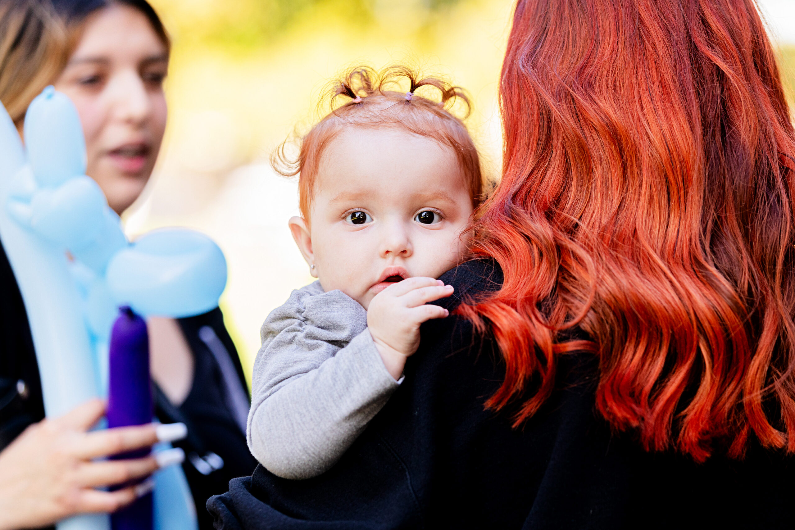 A baby with curly hair looks at the camera while being carried by an adult with long red hair. Another person holding a blue balloon animal stands nearby, slightly out of focus.