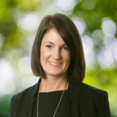 A woman with straight, shoulder-length brown hair, wearing a black blazer and top, is smiling in front of a blurred green and white outdoor background.