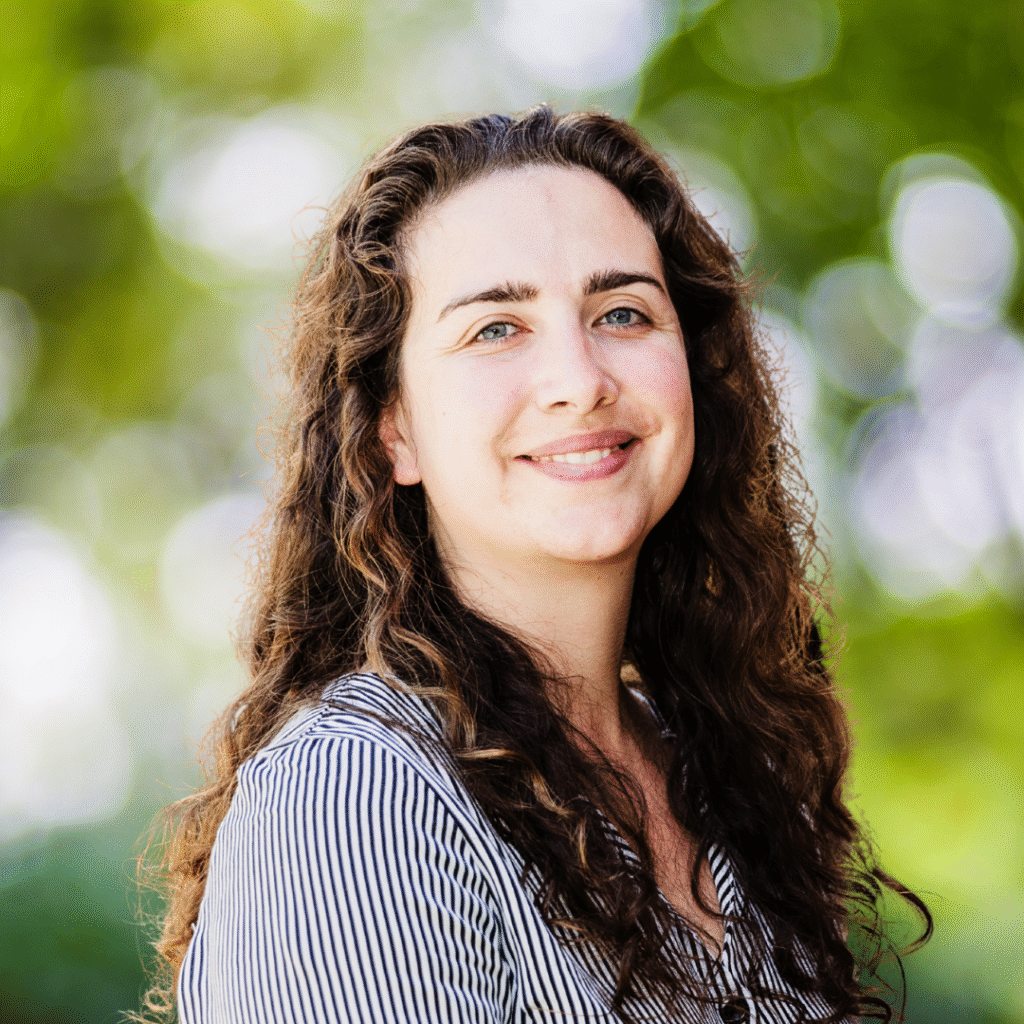 A woman with long curly brown hair and a striped shirt smiles outdoors, standing in front of a background of green trees and sunlight.