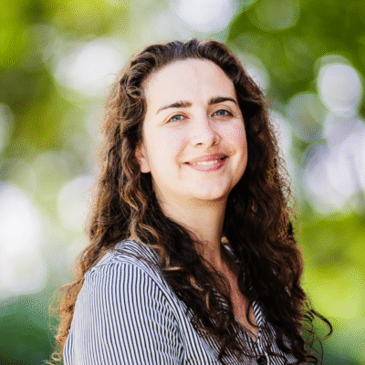 A woman with long curly brown hair and a striped shirt smiles outdoors, standing in front of a background of green trees and sunlight.