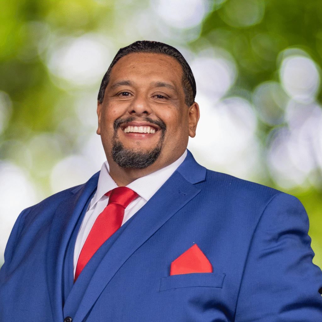 A man wearing a blue suit, white shirt, and red tie smiles confidently in front of a blurred green and white outdoor background.
