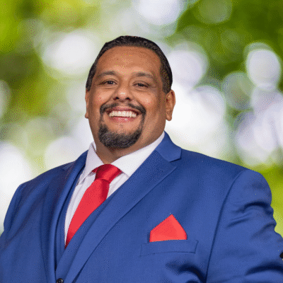 A man wearing a blue suit, white shirt, and red tie smiles confidently in front of a blurred green and white outdoor background.