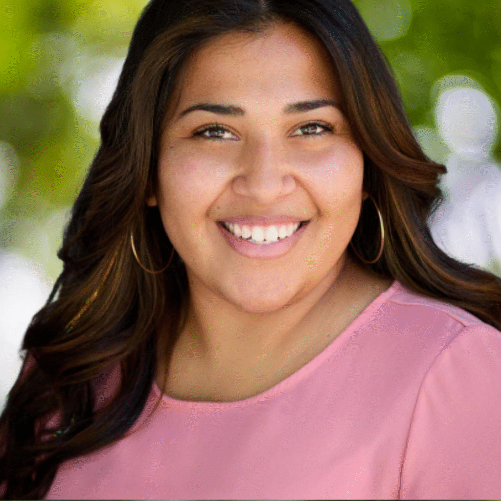 A woman with long brown hair and hoop earrings smiles at the camera. She is wearing a light pink top and is outdoors with a blurred green background.