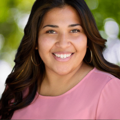 A woman with long brown hair and hoop earrings smiles at the camera. She is wearing a light pink top and is outdoors with a blurred green background.