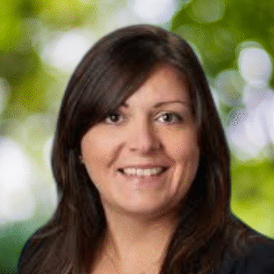 A woman with straight, dark brown hair and wearing a black top smiles at the camera. The background is blurred with shades of green and white, suggesting an outdoor setting with trees.
