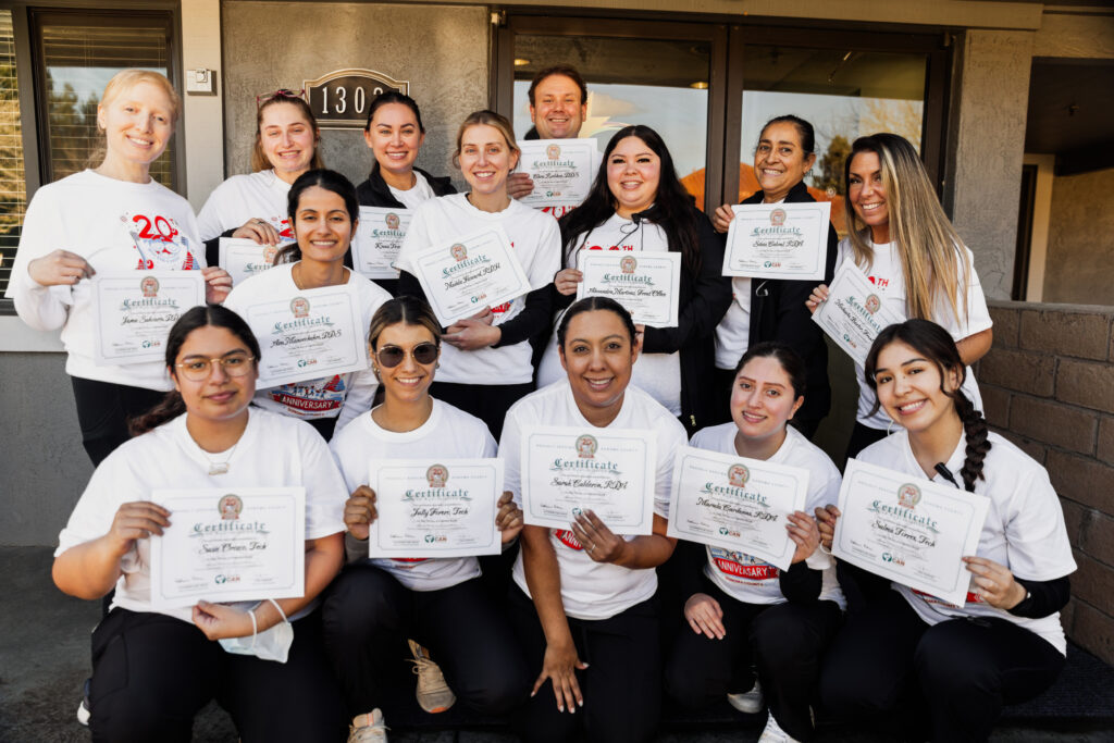 A group of thirteen smiling people pose outside, each holding a certificate. They wear matching white shirts, and appear happy and celebratory, suggesting they have completed a course or event together.