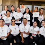 A group of thirteen smiling people pose outside, each holding a certificate. They wear matching white shirts, and appear happy and celebratory, suggesting they have completed a course or event together.