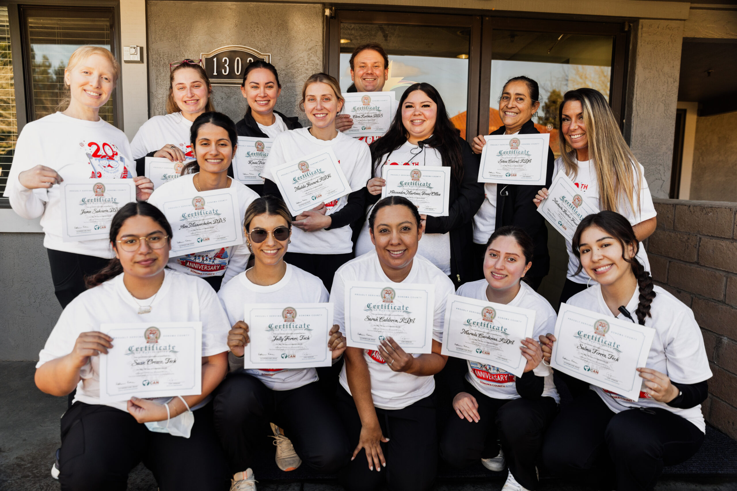 A group of thirteen smiling people pose outside, each holding a certificate. They wear matching white shirts, and appear happy and celebratory, suggesting they have completed a course or event together.