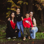 Four people, likely a family, pose outdoors in front of tall trees with autumn leaves. Two children stand in the middle, flanked by two adults, one squatting and one kneeling. Everyone is smiling except the girl in black.