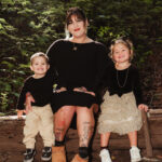 A woman sits on a log outdoors, smiling, with a young boy on her left and a young girl on her right. The children are both smiling. The background is lush greenery and dappled sunlight.