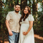 A couple stands together in a forest. The woman, wearing a white dress and holding her pregnant belly, smiles at the camera. The man, in a beige shirt and jeans, stands close beside her among tall trees and sunlight.