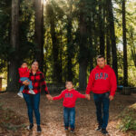 A family of four stands in a sunlit forest clearing, all wearing red tops. The parents hold hands with their two children, and tall trees surround them with sunlight streaming through the branches.