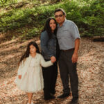 A family of three stands on a leaf-covered path in a wooded area. The smiling woman and man stand close together, holding hands with a young girl in a white dress and sweater. Sunlight filters through green trees in the background.