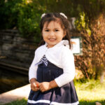 A young girl with dark hair in pigtails stands outdoors, smiling. She wears a navy blue dress with a white sweater and holds something small in her hands. Sunlight and greenery fill the background.