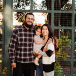 A smiling family of three poses outdoors by a wooden trellis. The man wears glasses and a plaid shirt, the woman wears a striped cardigan, and they are holding a young girl in a floral dress. Sunlight filters through the trees.