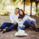 A man, woman, and young boy sit closely together on a brick path in a garden. All wear white shirts and jeans. A white cowboy hat sits on the ground in front of them. They look relaxed and smile softly at the camera.