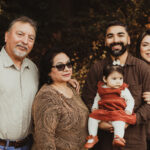 A family of five poses outdoors, smiling. An older man and woman stand next to a younger man and woman, who holds a baby girl in a red dress. Trees with autumn leaves are in the background.