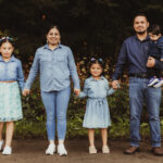 A family of five stands outdoors in front of greenery, holding hands. The parents and three children are dressed in blue denim outfits, with the father holding the youngest child. They are all smiling.