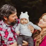 A man and woman smile at a baby wearing a white knit hat with bear ears. The man holds the baby while the woman looks on lovingly. They are outdoors with trees in the background.