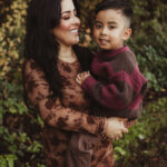 A woman in a brown floral dress holds a young boy dressed in a maroon and brown sweater. They are outdoors, smiling at each other, with green foliage in the background.