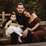 A man sits on a wooden bench holding a baby girl in a white dress with a green top. A woman in a brown dress sits beside him, resting her head on his shoulder. They are outdoors among trees, smiling at the camera.