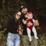 A smiling couple stands outdoors, holding a small black dog and a baby girl in a red outfit with a matching bow headband. The background is filled with greenery and trees.