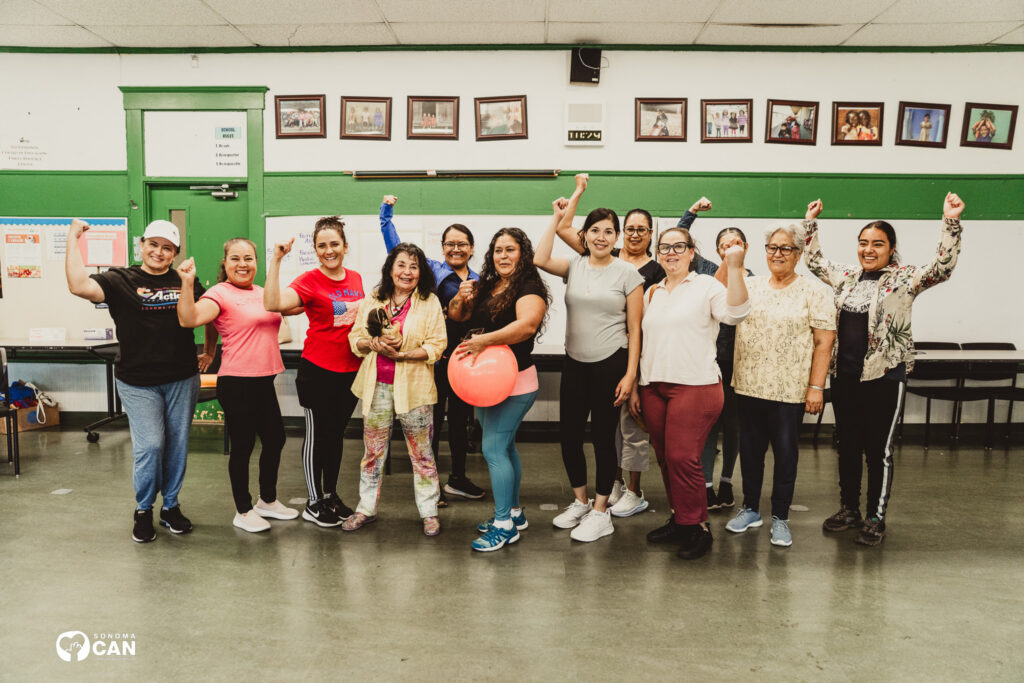 A group of women of various ages stand indoors, smiling and raising their fists in celebration. One holds a pink ball. The setting appears to be a community center with framed photos and posters on the walls.
