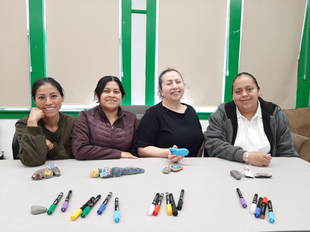 Four women sit at a table displaying painted rocks and markers. They are smiling, and the table in front of them holds various decorated stones and colorful markers. Beige blinds and green window frames are in the background.