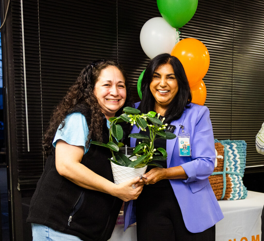 Three smiling adults stand indoors at an event. Two women hold a potted plant together, while a man in the background holds a bowl. Colorful balloons and a "Sonoma CAN" sign are visible behind them.